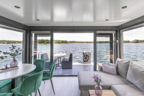 Un interior moderno con vista a un lago, un comedor con mesa y sillas, y una zona de estar con sofá y cojines.