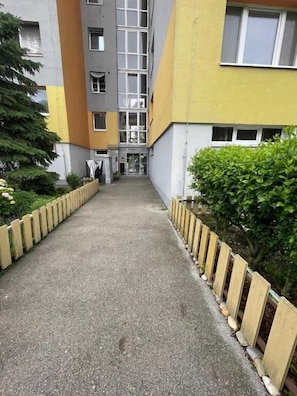 A pathway leading to a building with a yellow and gray facade, surrounded by greenery and a wooden fence.
