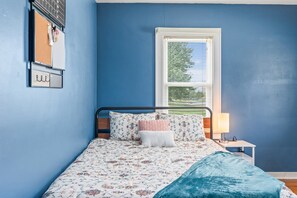 A bedroom with a bed, a nightstand, a lamp, a window, and a wall-mounted shelf. - Roadmap Lodging: 3BR Near Columbus Regional Health (Columbus)