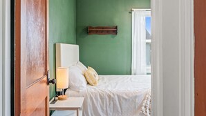 A bedroom with a green wall, a wooden bed frame, a bedside table with a lamp, and a window with white curtains. - Roadmap Lodging: Near Columbus Regional Health, 3BR Monthly, Pet OK (Columbus)