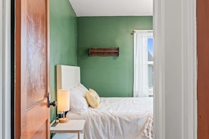 Ein Schlafzimmer mit grüner Wand, einem hölzernen Bettgestell, einem Nachttisch mit Lampe und einem Fenster mit weißen Vorhängen.