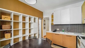 Cozinha com armários brancos, mesa de jantar e cadeiras. Há prateleiras com cestos e uma porta que dá para outro espaço.