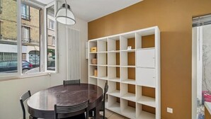 A room with a round table, chairs, a white shelving unit, and a window with a view of the street. - Charming Fully Equipped 1BR - Near Paris (Boulogne-Billancourt)
