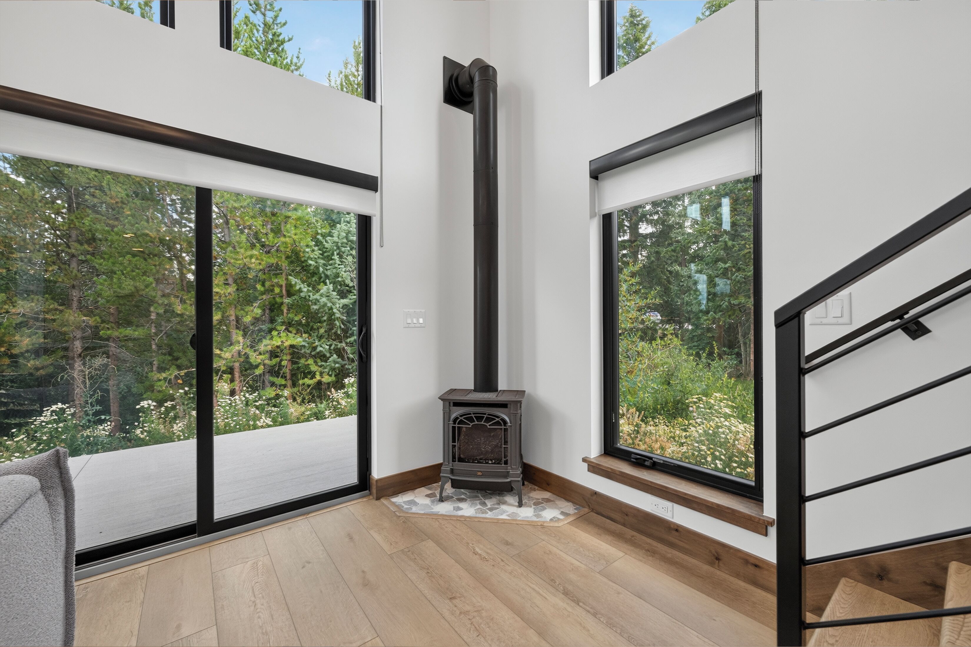 A modern interior with a wood-burning stove, large windows, and a staircase.