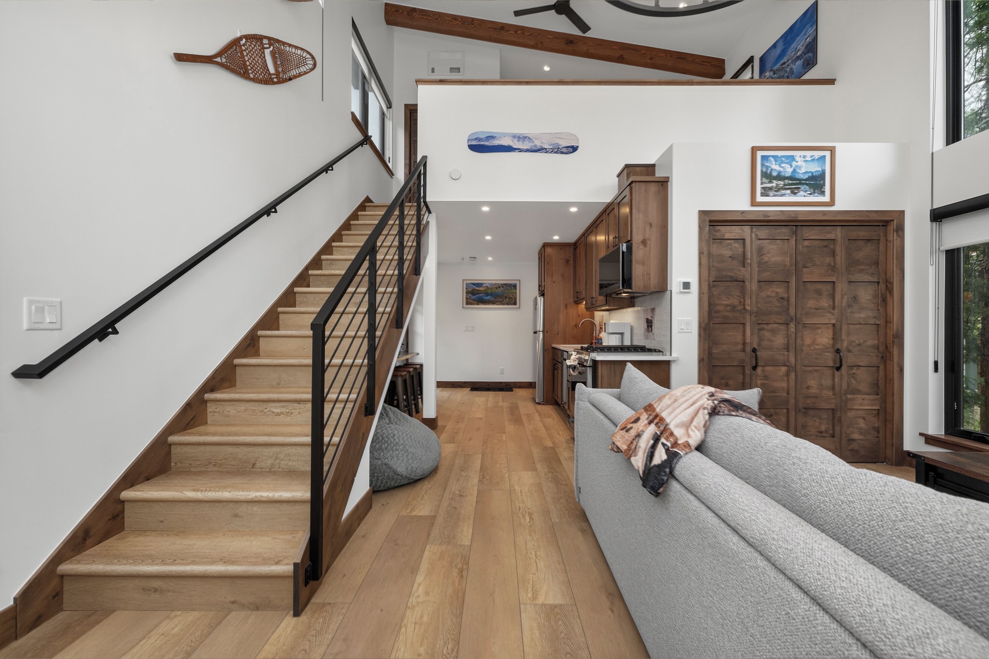 Un escalier avec des marches en bois et une rampe noire, menant à un espace de vie comprenant un canapé gris et une porte en bois.