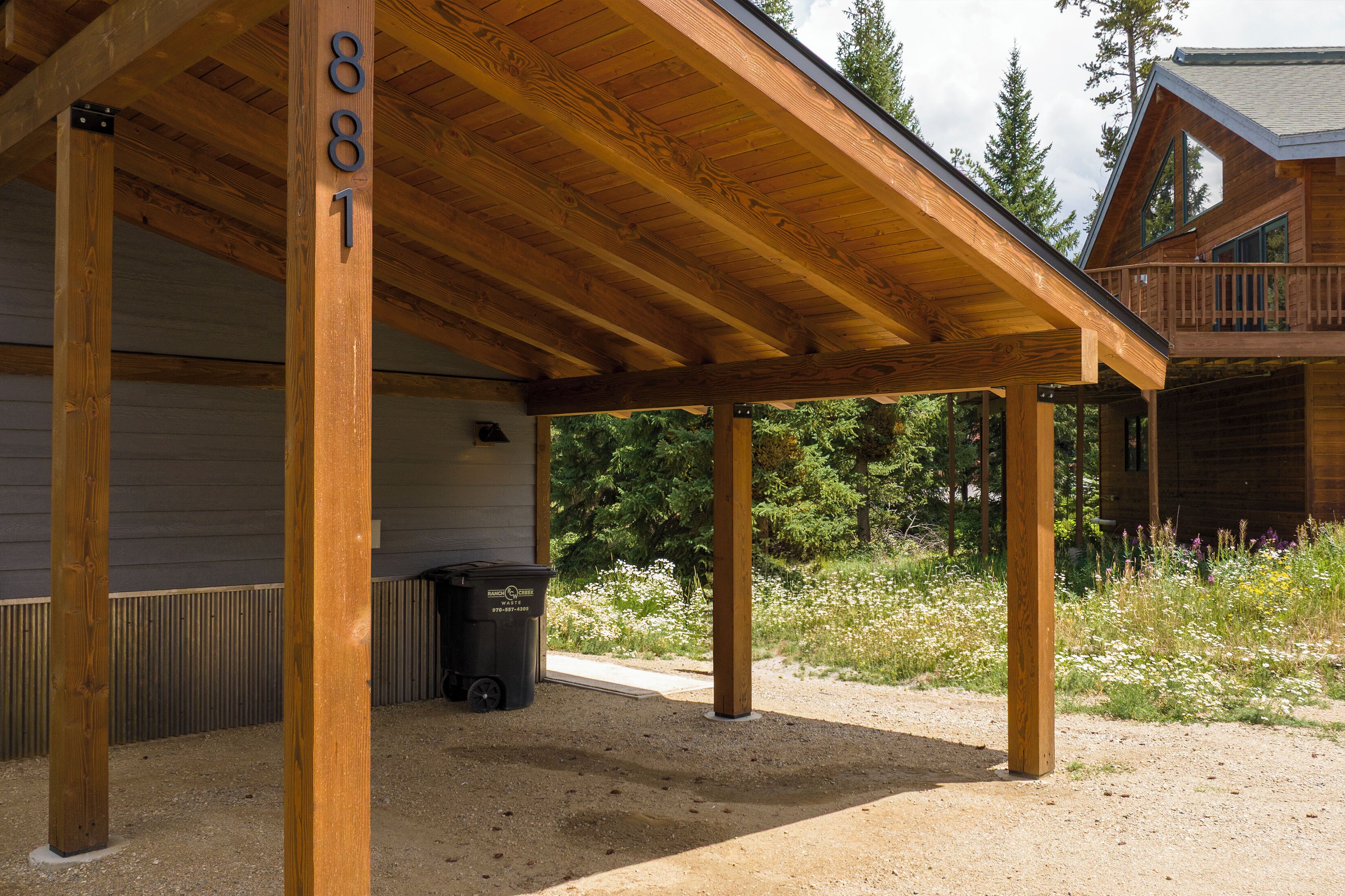 A covered outdoor area with wooden beams and a gravel ground, a trash bin, and a building in the background.