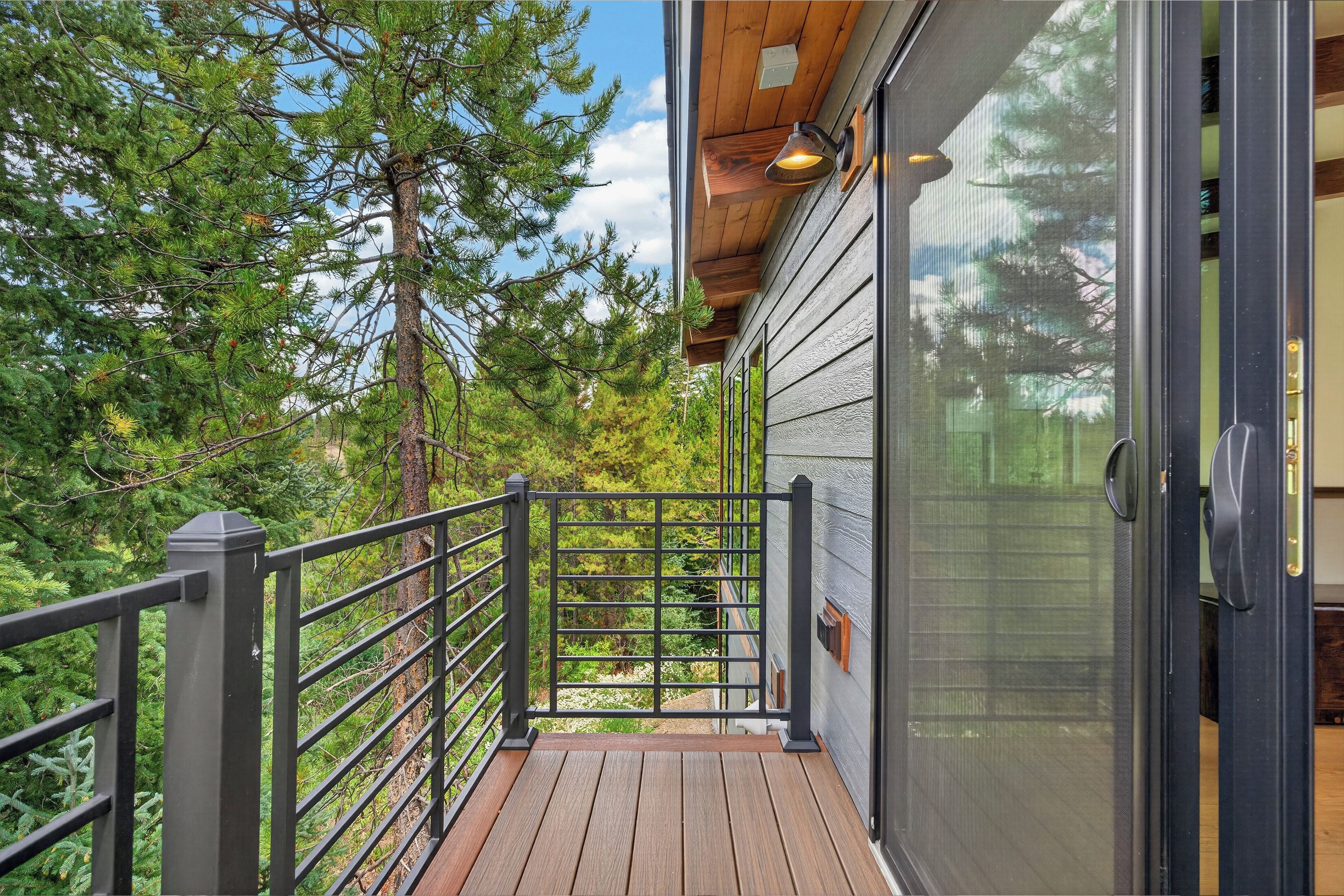 A wooden deck with a metal railing leading to a glass door, surrounded by trees.