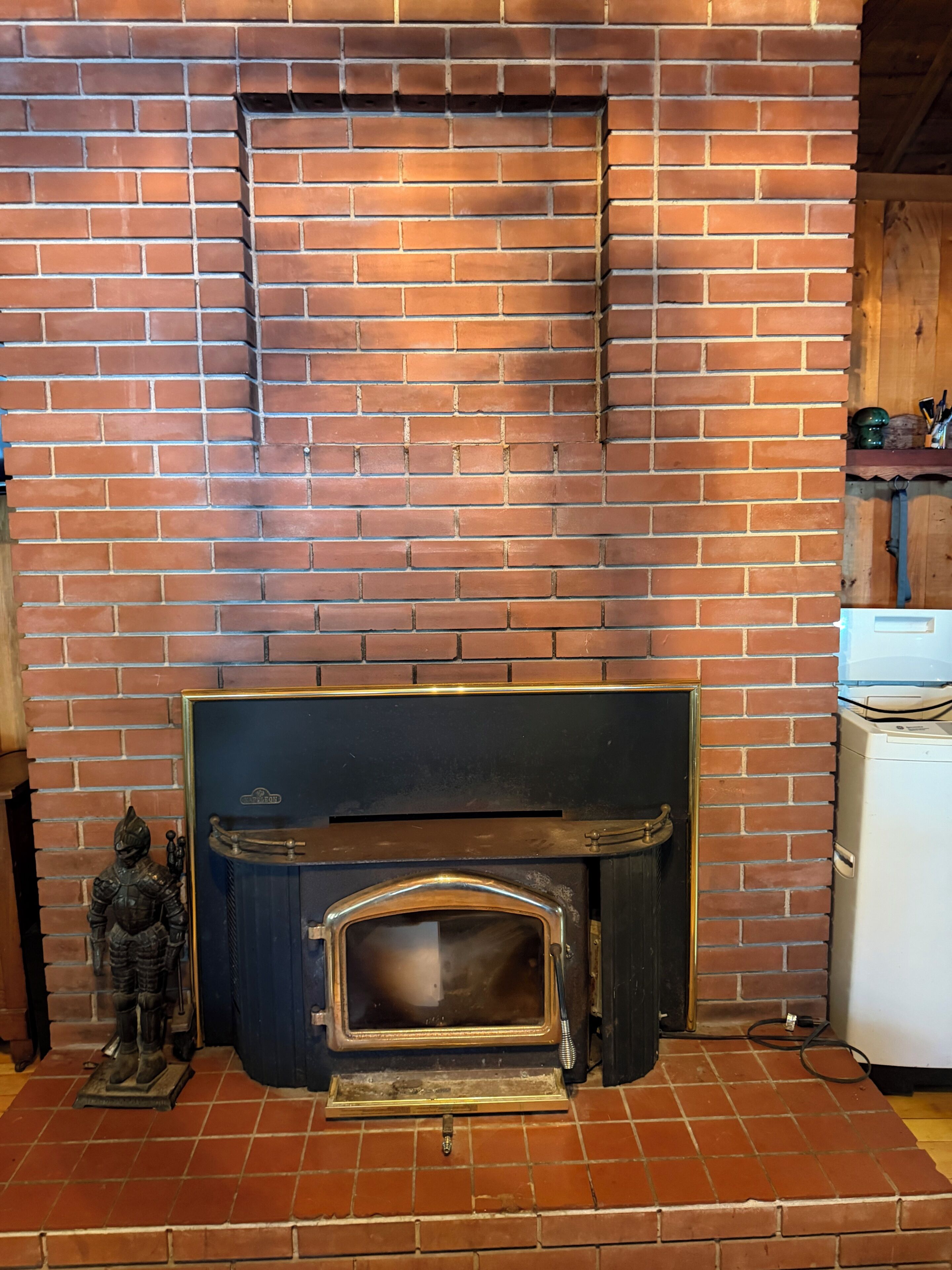 Una chimenea de ladrillo con estructura metálica, una pequeña estatua a la izquierda y un electrodoméstico blanco a la derecha.