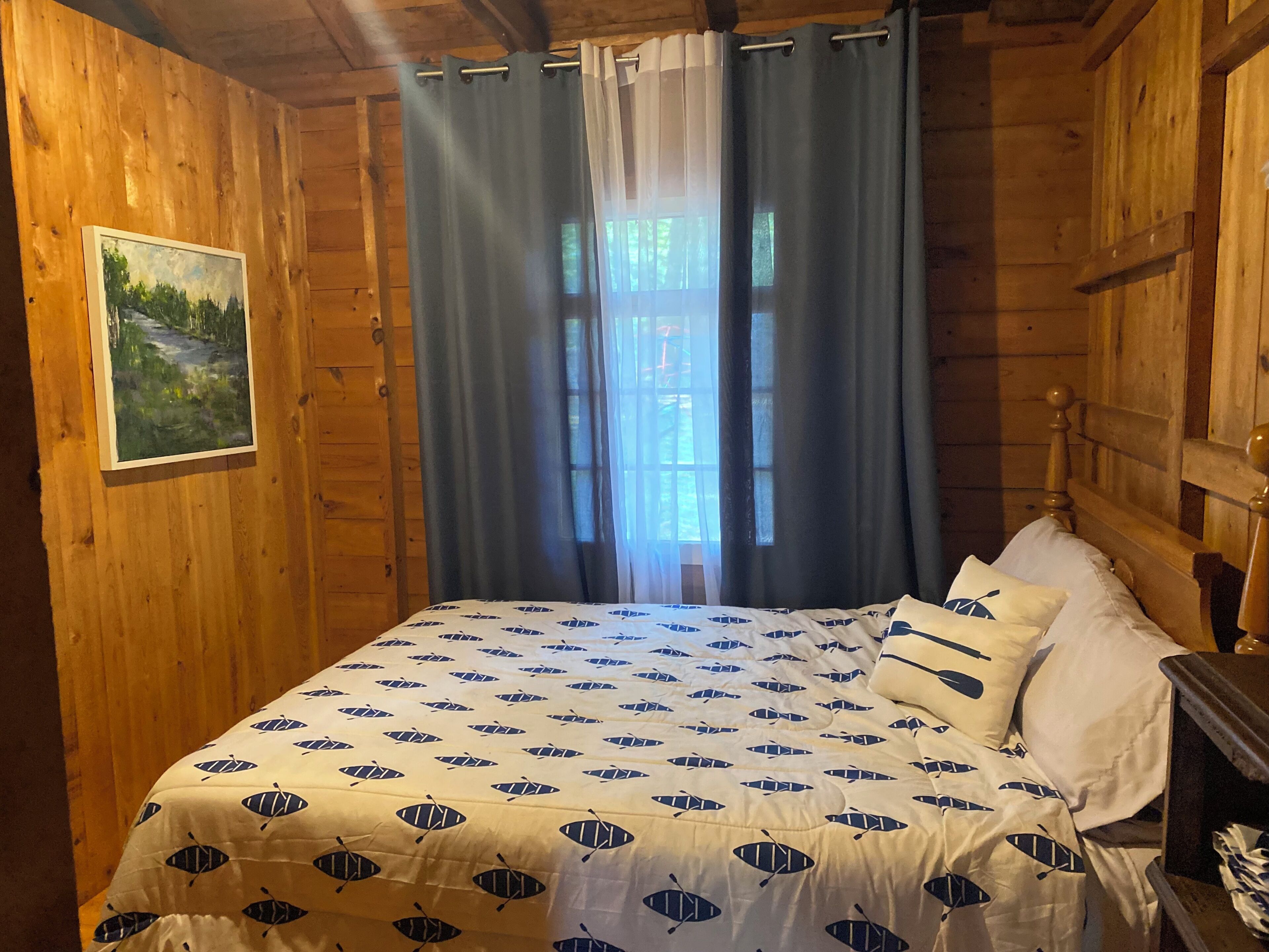 Una habitación de madera con cama, un cuadro en la pared y una ventana con cortinas.