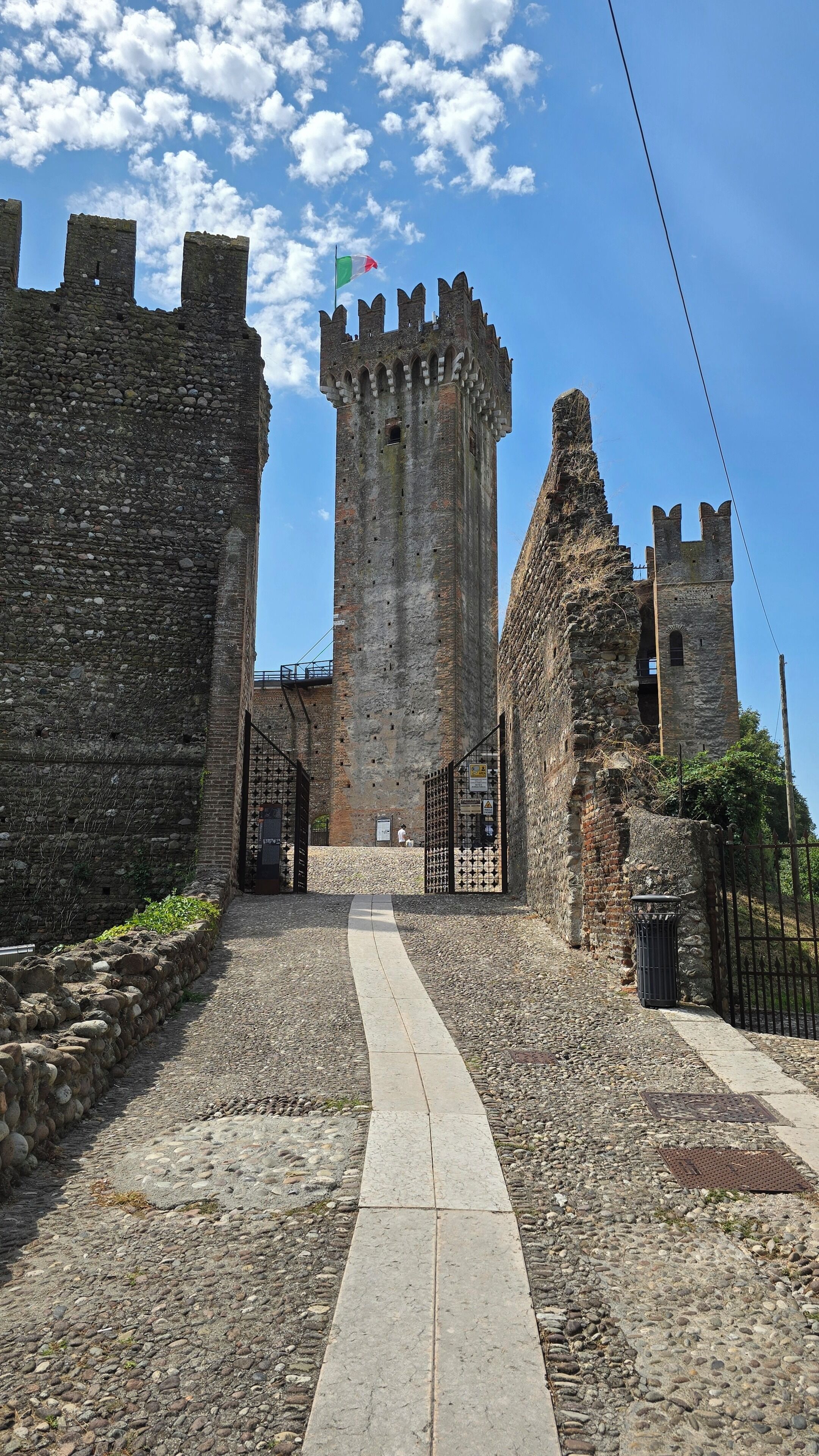 Una porta de pedra històrica que dona pas a un castell amb una bandera, un camí pavimentat i un cel serè.