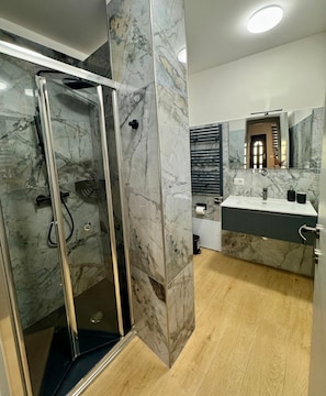 A modern bathroom with a glass shower enclosure, marble walls, a wooden floor, and a large mirror above the sink. - Casa Magenta Apartment 4 (Valeggio sul Mincio)