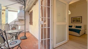 Un balcone con un tavolo e sedie in maiolica, una porta scorrevole in vetro che dà su una camera da letto con un letto e un quadro appeso al muro.