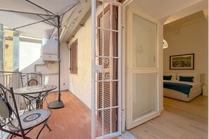 Ein Balkon mit einem gefliesten Tisch und Stühlen, eine Glasschiebetür, die zu einem Schlafzimmer mit einem Bett und einem Bild an der Wand führt.
