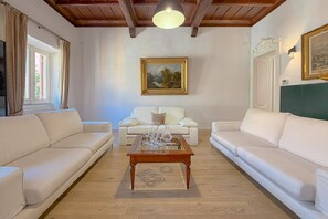 Uma sala de estar com dois sofás brancos, uma mesa de centro de madeira com copos, um quadro na parede e uma janela com cortinas.