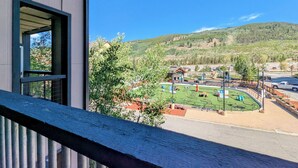Udsigt fra en balkon over et legeområde med en grøn mark, en bygning med saddeltag og en skovklædt bakke i baggrunden.