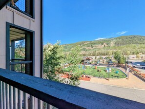 Blick vom Balkon auf einen Spielplatz mit einer grünen Wiese, ein Gebäude mit Satteldach und ein bewaldeter Hügel im Hintergrund.