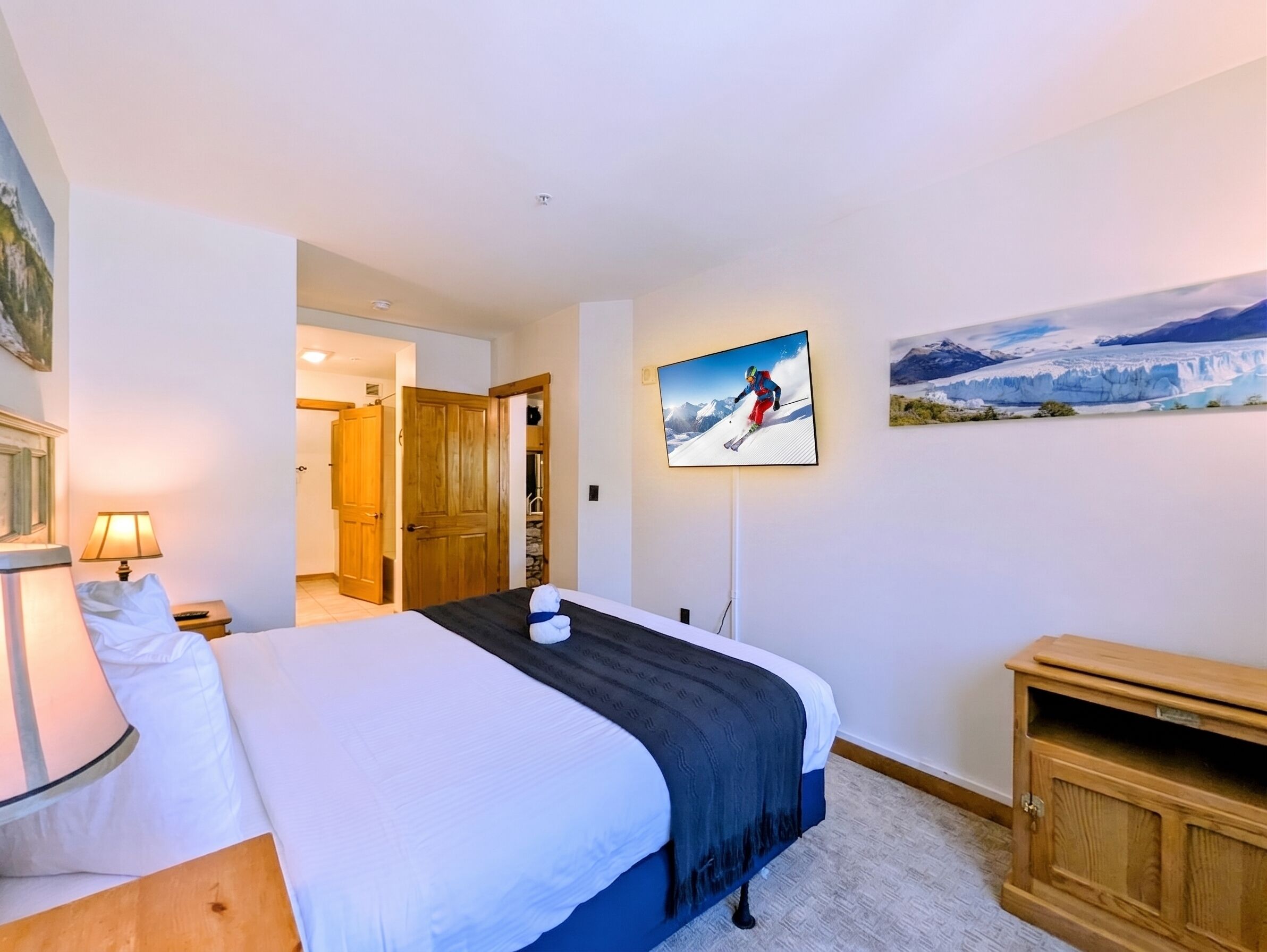 A hotel room with a bed, a wooden cabinet, a painting of a skier, and a painting of a glacier.