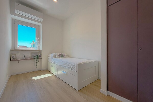 Ein Schlafzimmer mit einem Einzelbett, einem Fenster mit Ausblick, einem Kleiderschrank und einem Regal mit Dekoration.