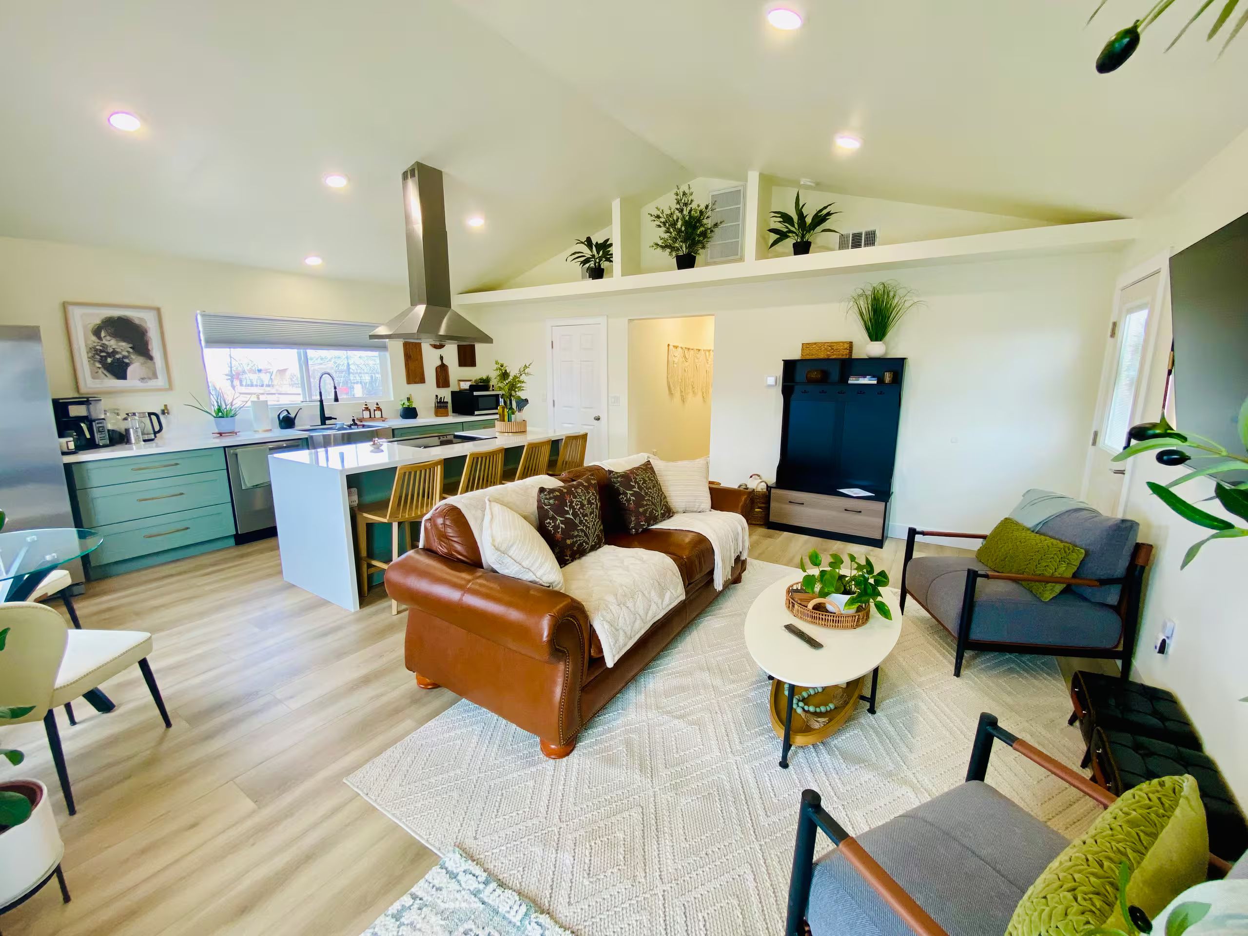 A modern living space with a brown leather sofa, a dining area with wooden chairs, a kitchen with a central island, and a TV mounted on the wall.