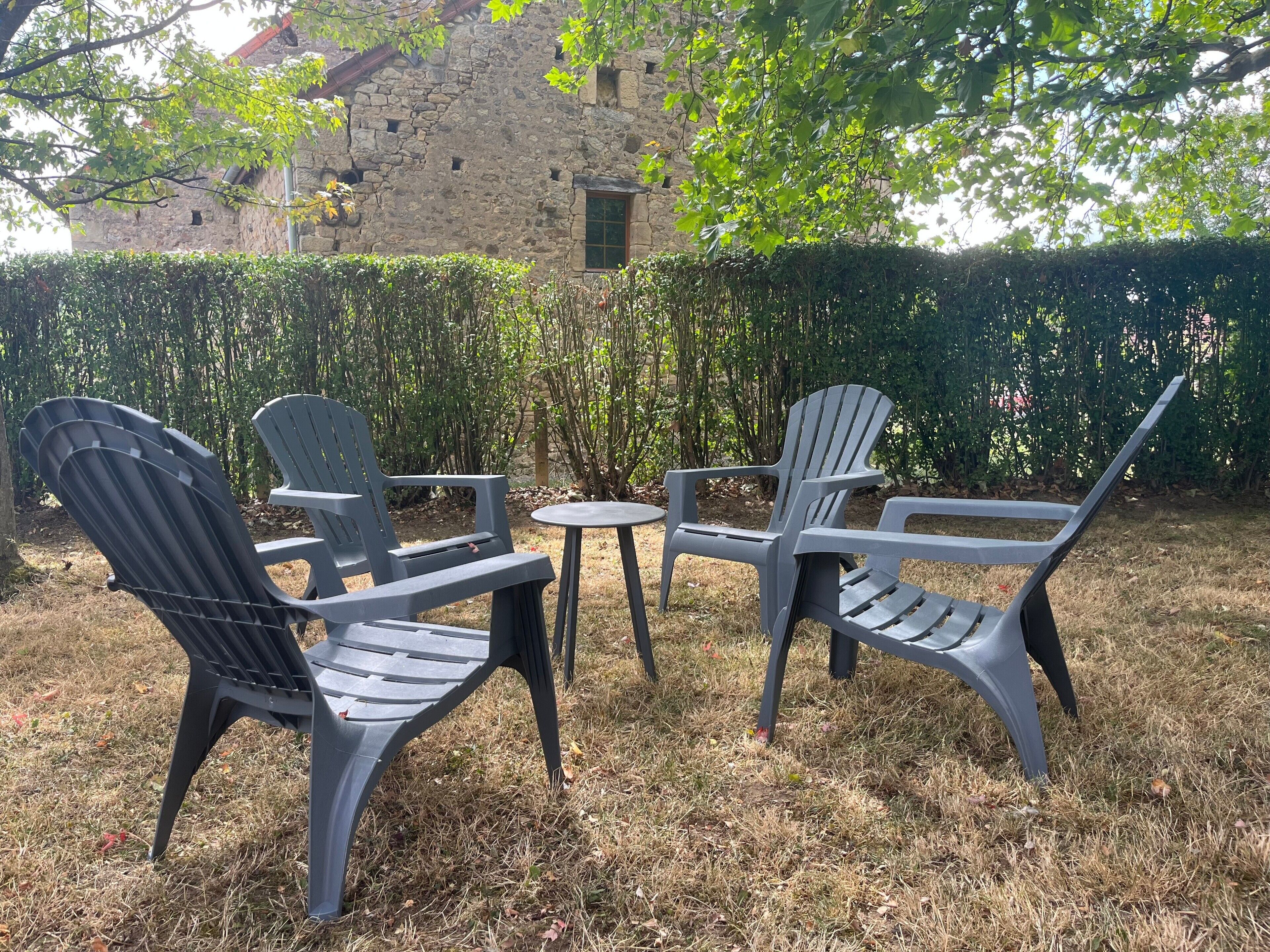 Un espacio al aire libre con dos sillas y una mesa pequeña, rodeado de un seto y un edificio de piedra al fondo.
