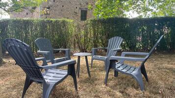 Un espacio al aire libre con dos sillas y una mesa pequeña, rodeado de un seto y un edificio de piedra al fondo.
