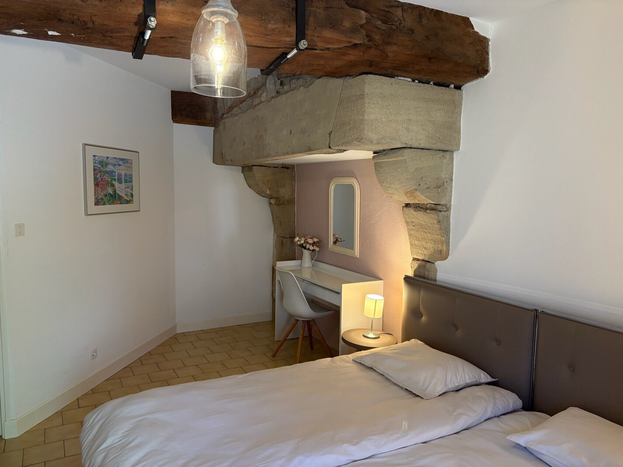 A bedroom with a bed, a desk, and a chair, a mirror, and a hanging light fixture.