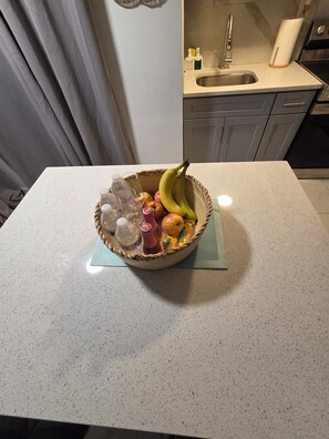 A kitchen counter with a fruit bowl containing bananas, apples, and a bottle. - Tropical Oasis: Cozy 1- bedroom with AC near JFK Airport, LIRR Train (Queens)