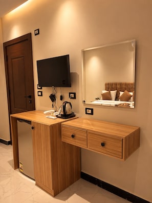 A hotel room with a wooden desk, a flat-screen TV mounted on the wall, a bed with a headboard, and a mirror reflecting the bed.