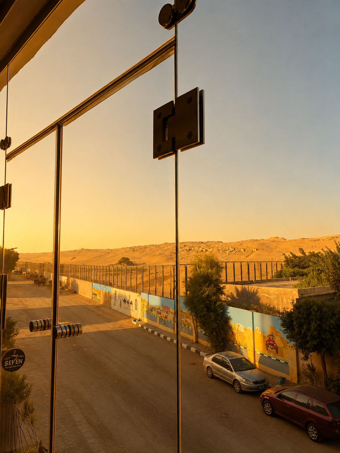 A view through a window with a metal structure, a road with cars, and a distant landscape with hills.