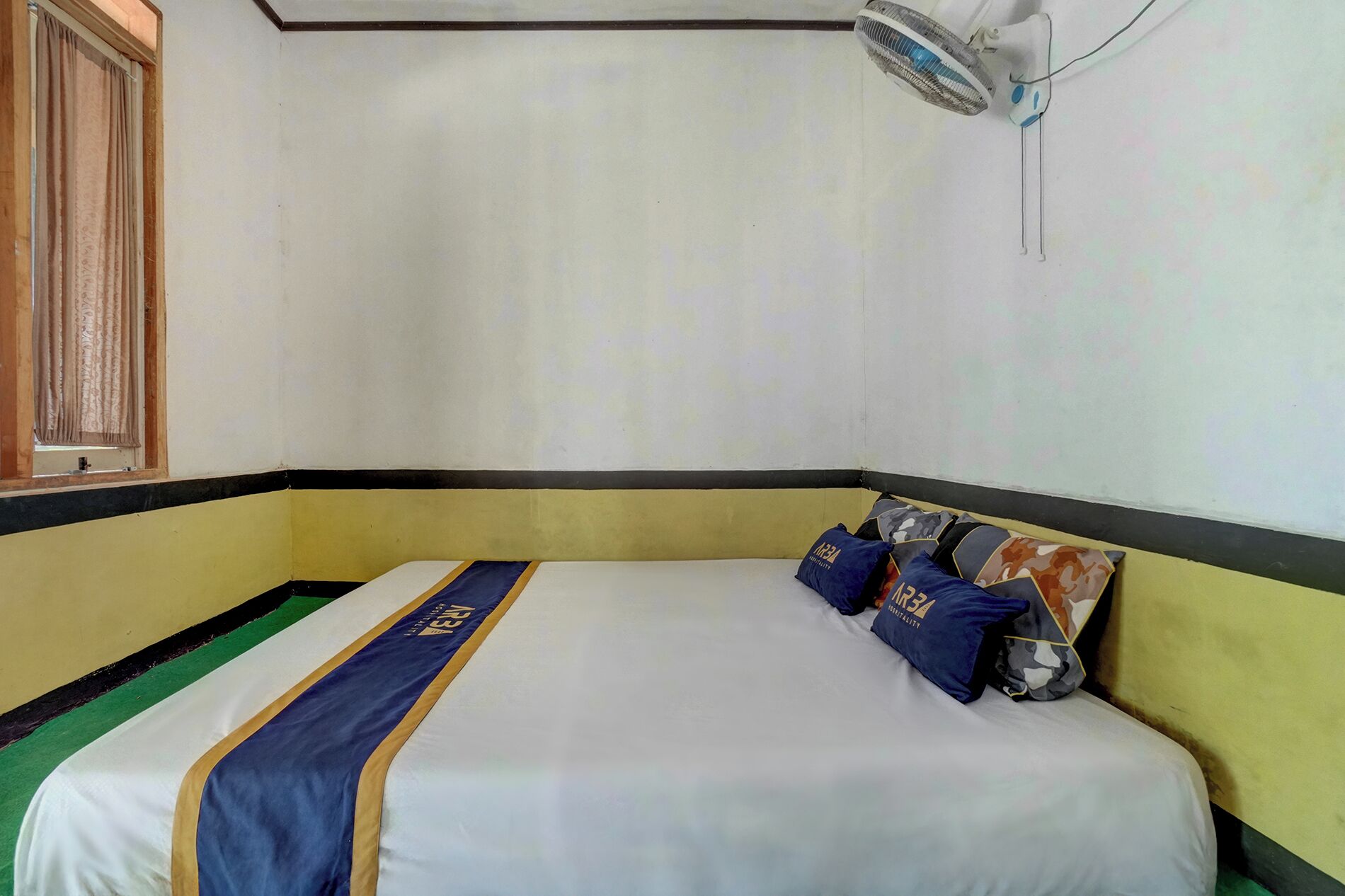 A single bed with a striped bedspread and pillows, a window with curtains, and a wall-mounted fan.