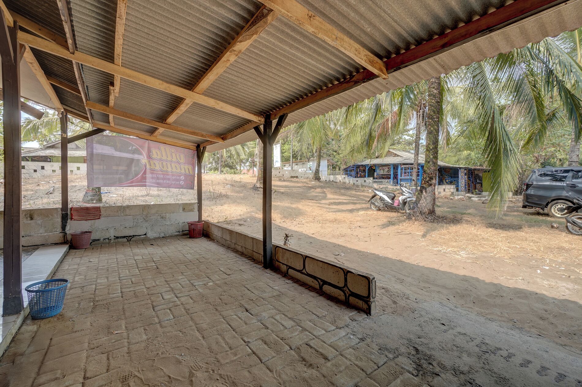 A covered outdoor area with a brick floor, a metal roof, and a view of a rural setting with trees and a parked car.