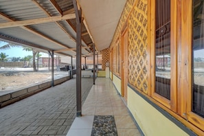 A covered outdoor area with a tiled floor, wooden columns, and a yellow wall with patterned windows.