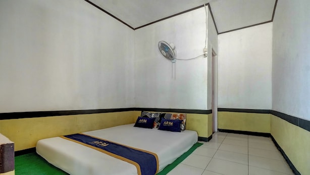 Una cama individual con colchón blanco, colcha azul y dorada, y dos almohadas con un logotipo. Un ventilador pequeño está montado en la pared sobre la cama.