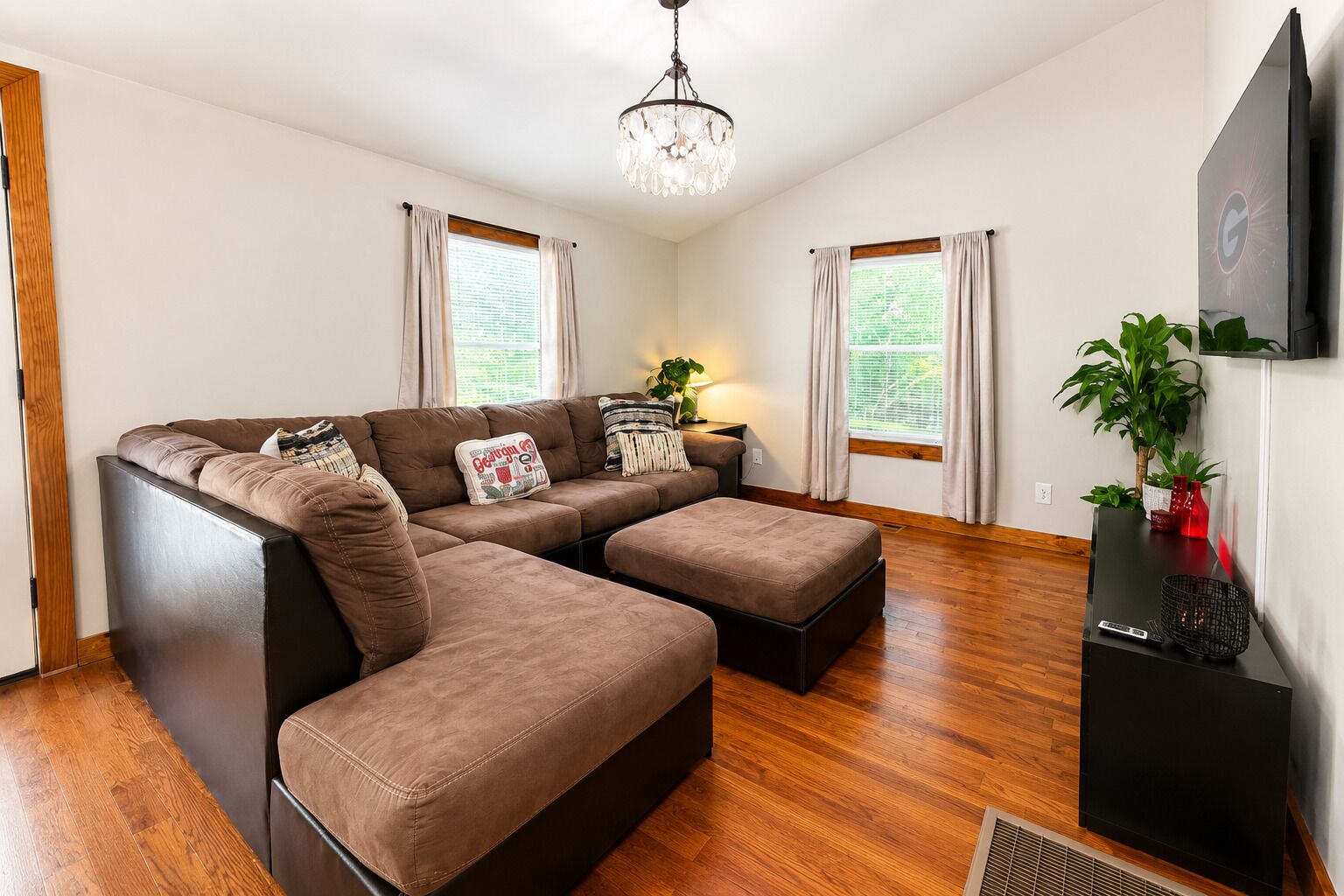 A modern living room with a sectional sofa, a small table, a chandelier, and a flat-screen TV.