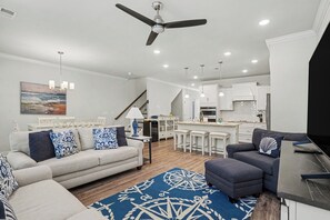 Ein modernes Wohnzimmer mit Sofa, Sesseln, einem Couchtisch, einem Essbereich und einer dahinterliegenden Küche sowie einem Deckenventilator.