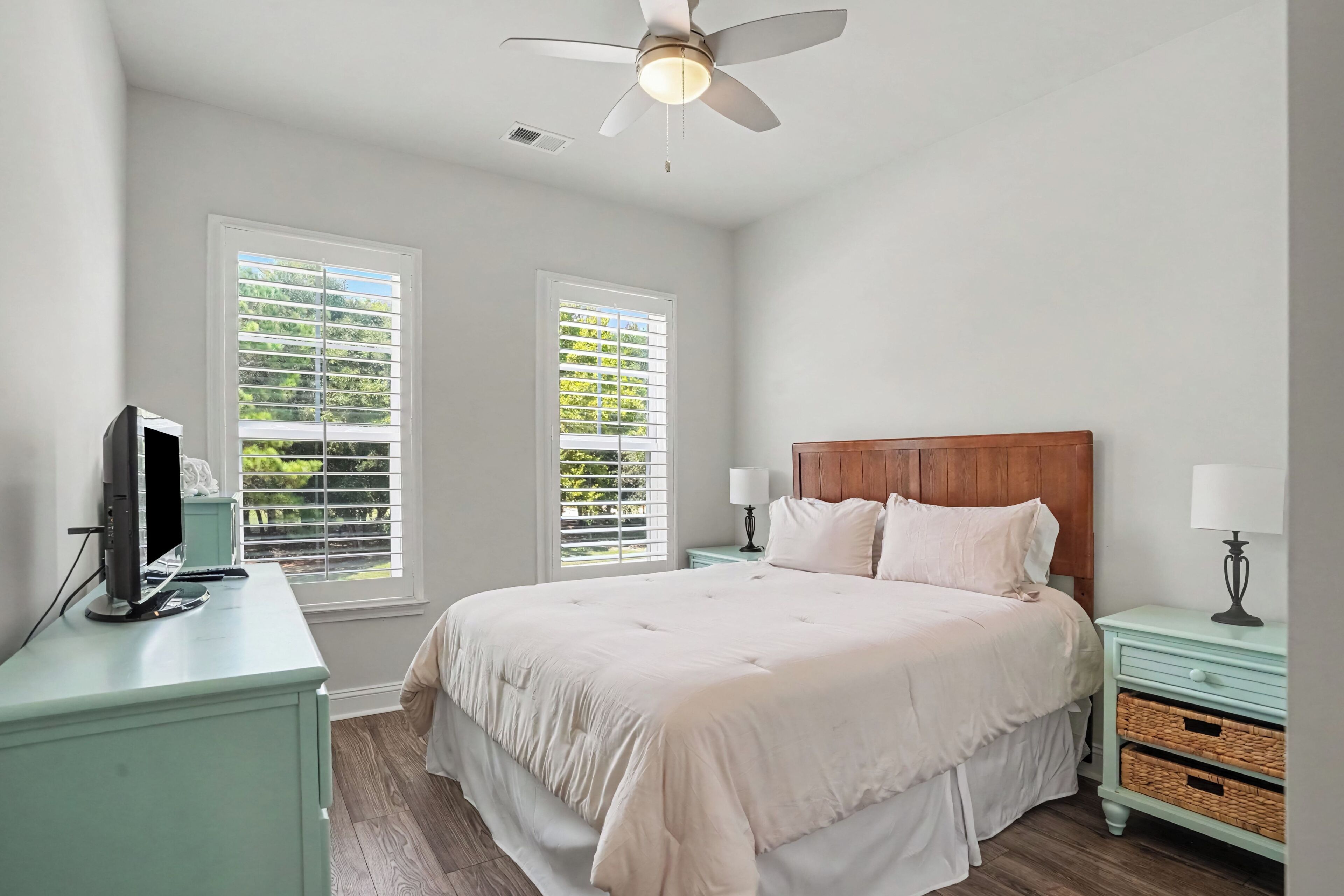 A bedroom with a bed, a desk with a computer, two windows with blinds, a ceiling fan, and a nightstand with a lamp.