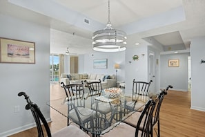 A dining area with a glass table surrounded by chairs, a chandelier, and a view of a living room with a sofa and a painting. - New Property On Program! North Tower #207 in Barefoot Resort! (North Myrtle Beach)