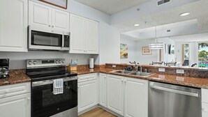 Una cocina moderna con armarios blancos, electrodomésticos de acero inoxidable, encimeras de granito y vistas al exterior.