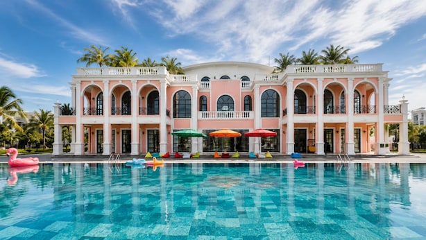 A large building with multiple arches and balconies, a swimming pool with colorful lounge chairs, and palm trees in the background. - Hoang Linh Hotel Phu Quoc (Phu Quoc)