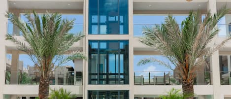 A modern building with a central glass elevator shaft, surrounded by palm trees and a swimming pool.
