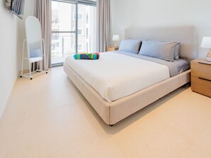 A modern hotel room with a large bed, a bedside table with a lamp, a small side table, and a window with curtains. - Modern Condo Near Eagle Beach-Balcony, Pool & Gym, Brand new Walishali Condo (Oranjestad)