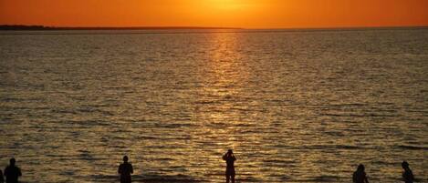 A sunset over a calm sea with silhouettes of people on the shore.
