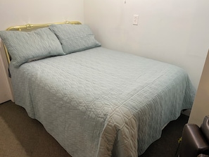 A single bed with a quilted headboard and footboard, a power outlet on the wall, and a brown leather chair in the corner.