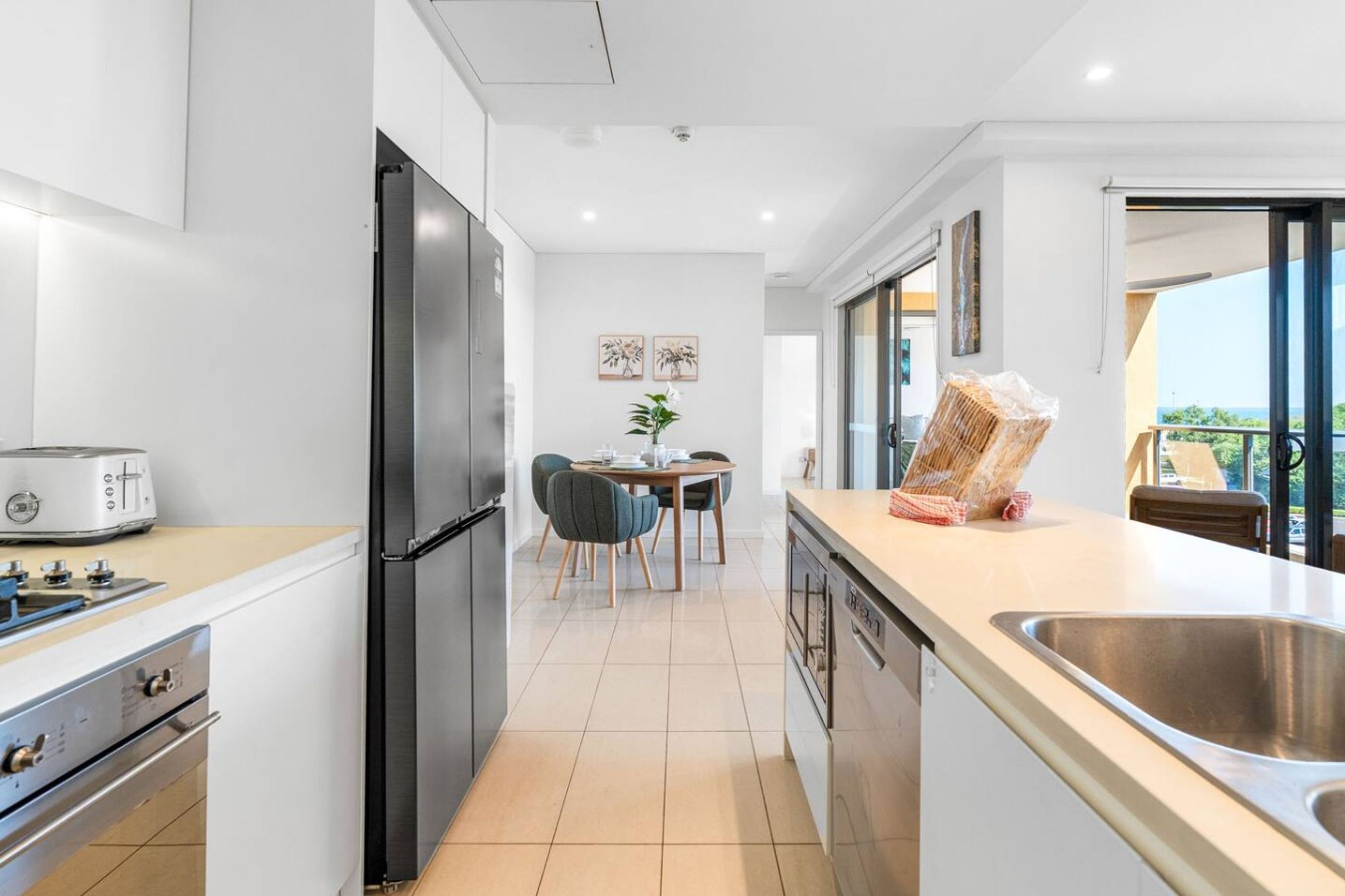 A modern kitchen with a refrigerator, stove, and dishwasher. There is a dining area with chairs and a table, and a balcony with a view.