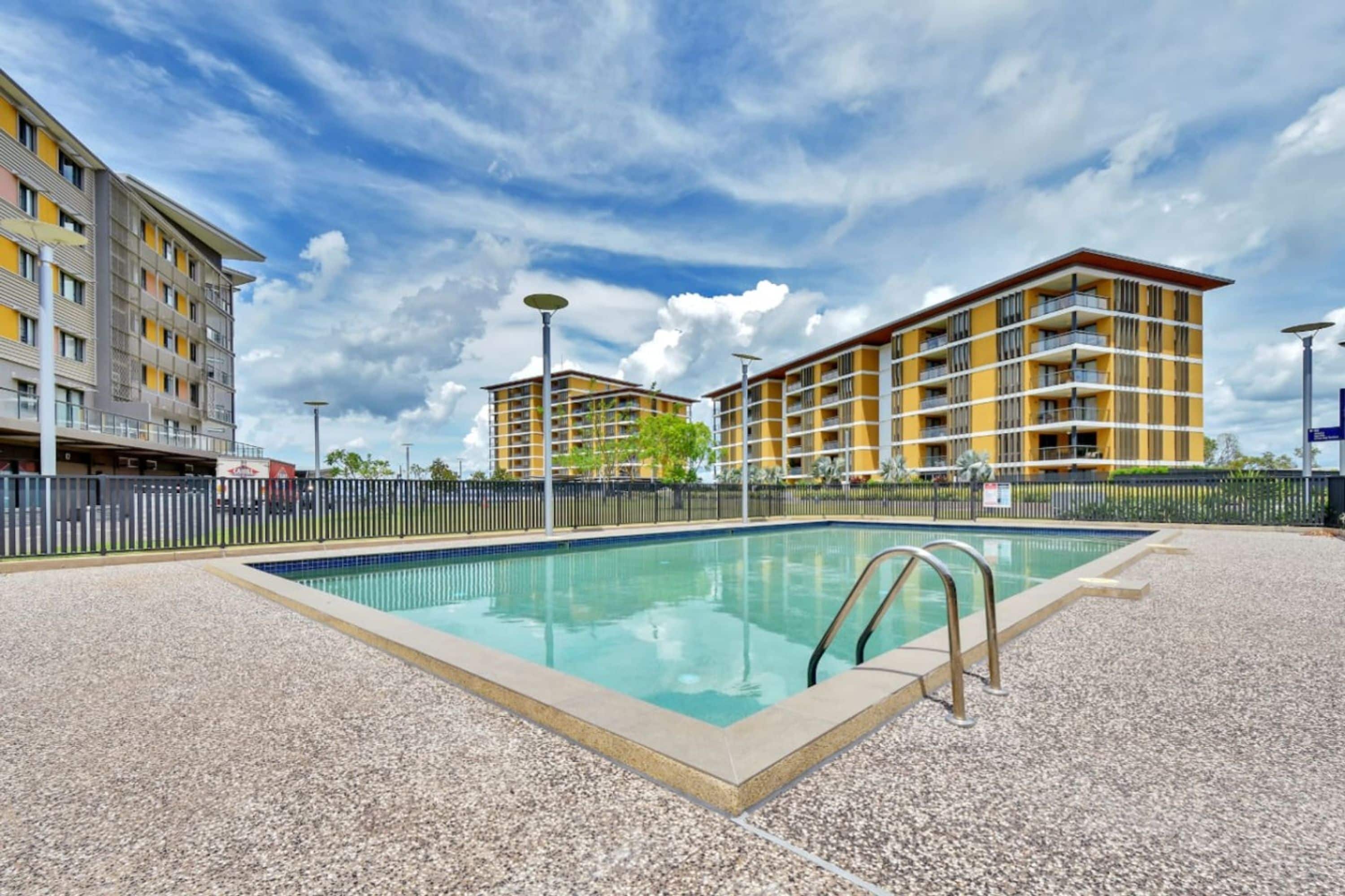A swimming pool with a metal ladder, surrounded by a gravel area, with multi-story buildings in the background.
