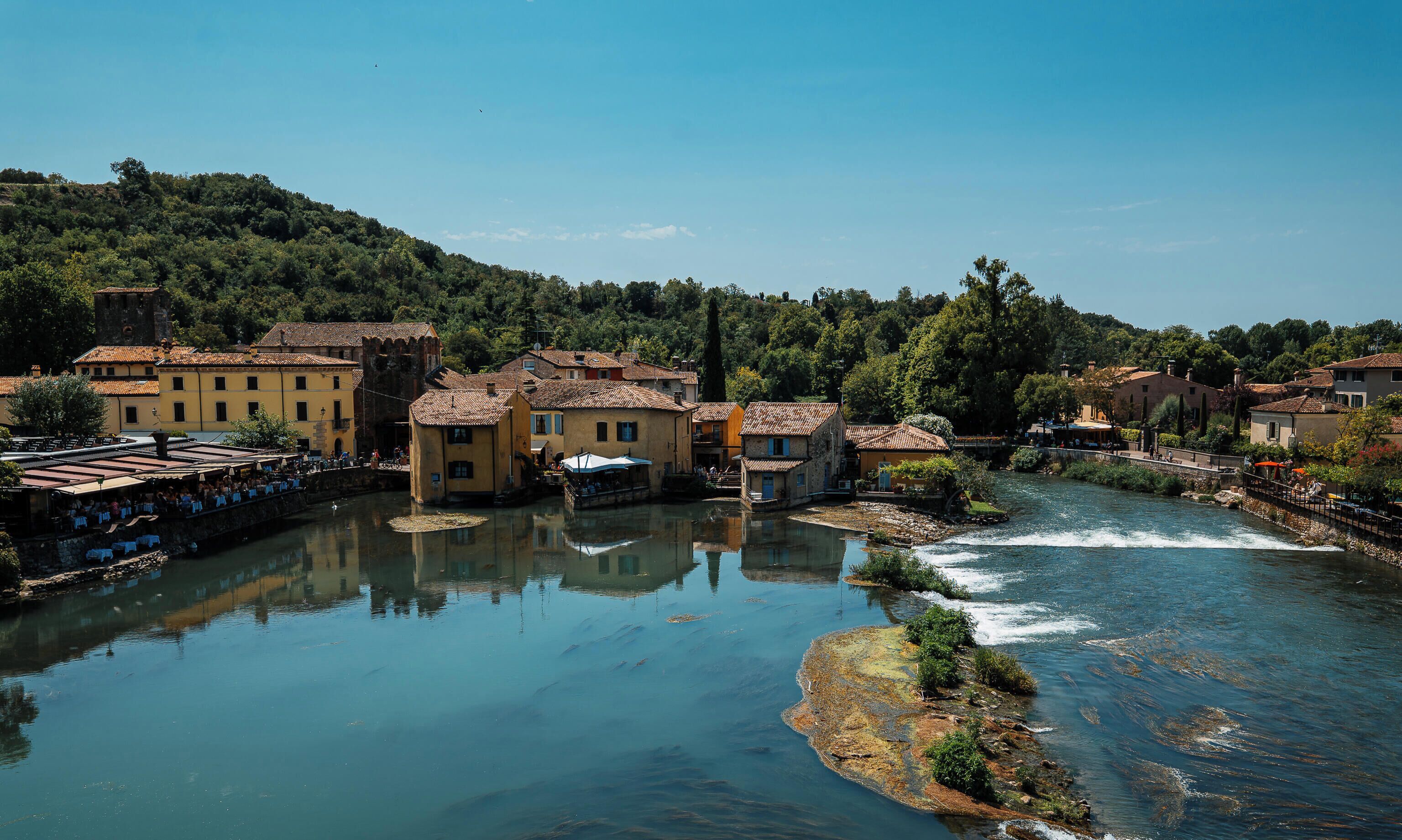A riverside town with buildings along the water, a bridge, and a hill in the background.