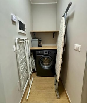A laundry room with a washing machine, dryer, ironing board, and a small shelf with a kettle. - Casa Magenta Apartment 5 (Valeggio sul Mincio)