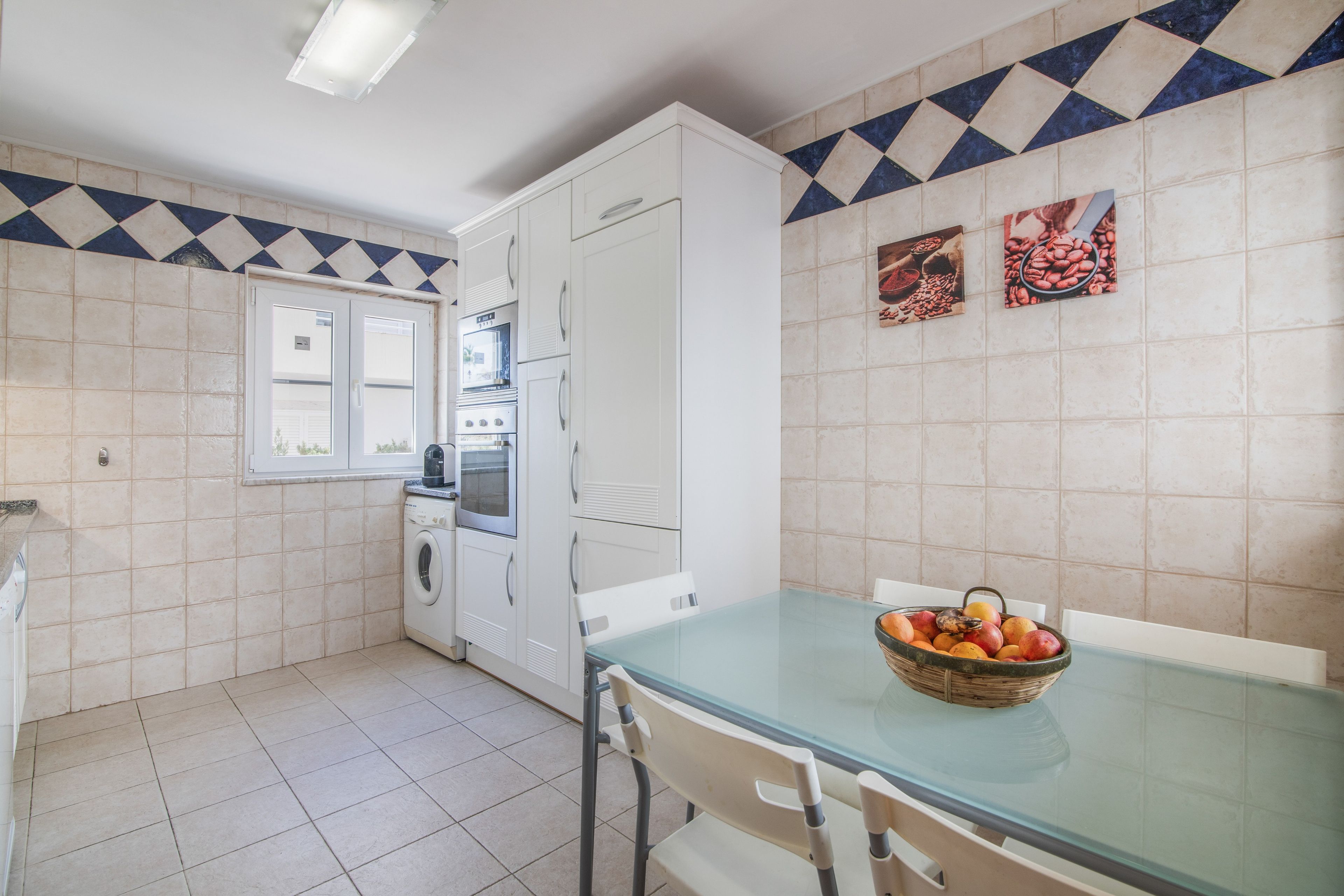 Une cuisine avec des meubles blancs, un four encastré, un lave-linge et une table à manger avec des chaises.