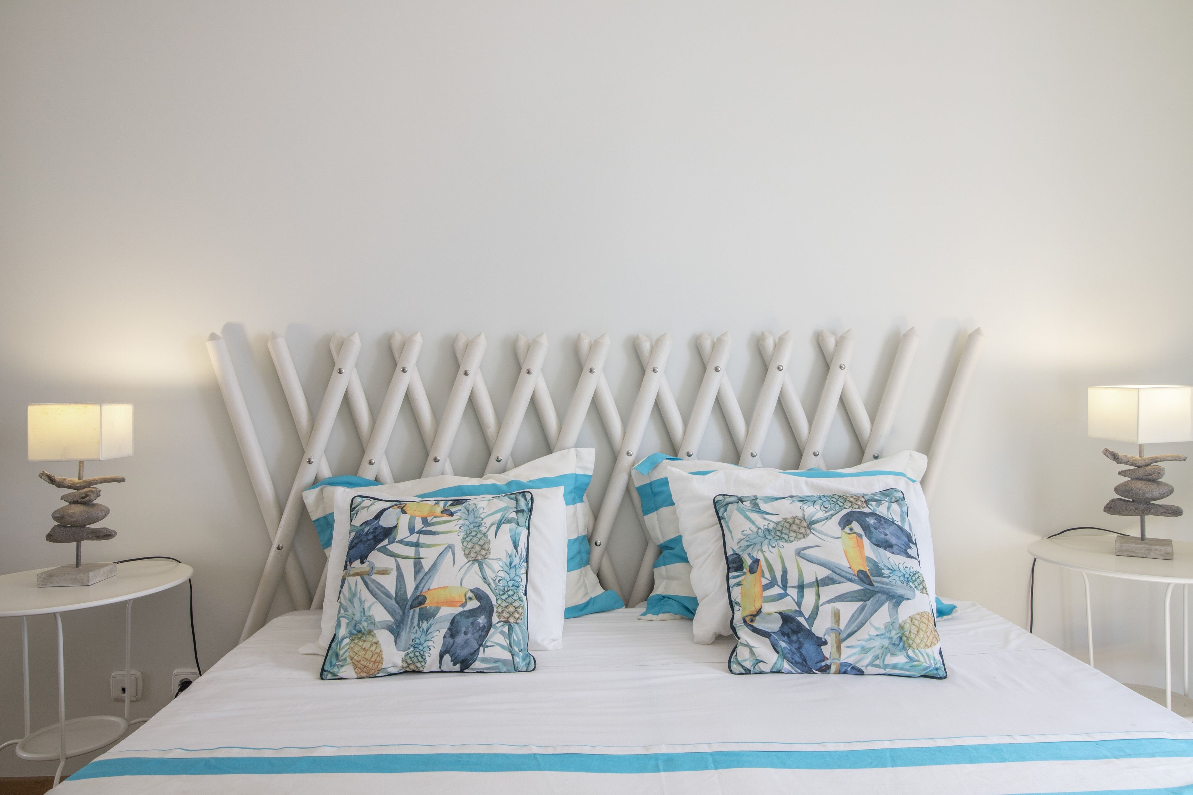 Un lit avec une tête de lit blanche, deux oreillers assortis à motifs tropicaux et deux lampes de chevet au design simple.