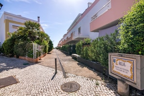 A paved pathway leading to a residential area with a sign indicating 'Rua' and 'TRISTÃO VAZ TEIXEIRA'.